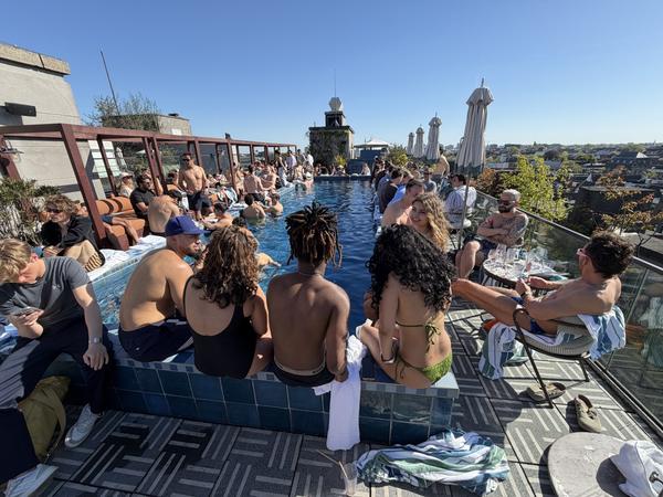 A vibrant rooftop pool party is in full swing under a clear blue sky, with many people enjoying the sun and water